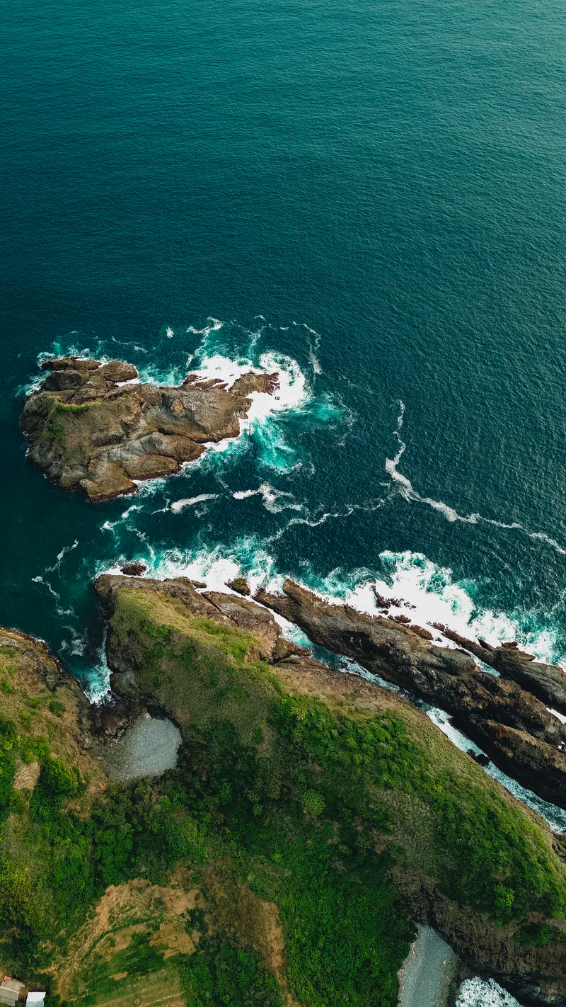 Surf breaking around rocks at the foot of a green headland, seen from above