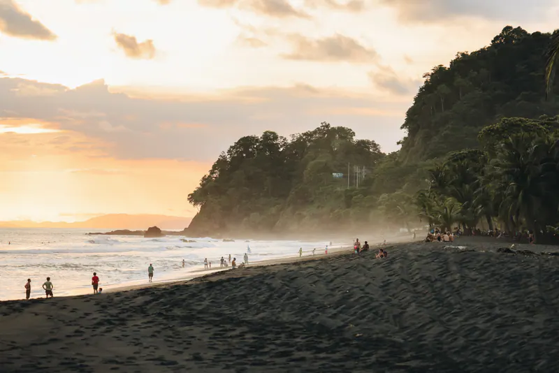 People walking along a dark sand beach at sunset, with a wooded headland beyond