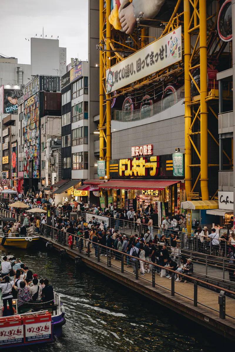 Crowds and a tour boat along a canal in Osaka, under bright signs