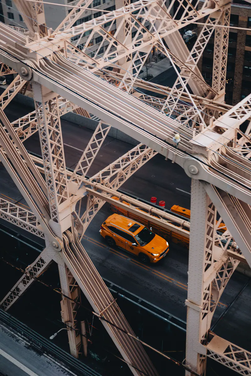 A yellow taxi seen through the steel beams of a bridge