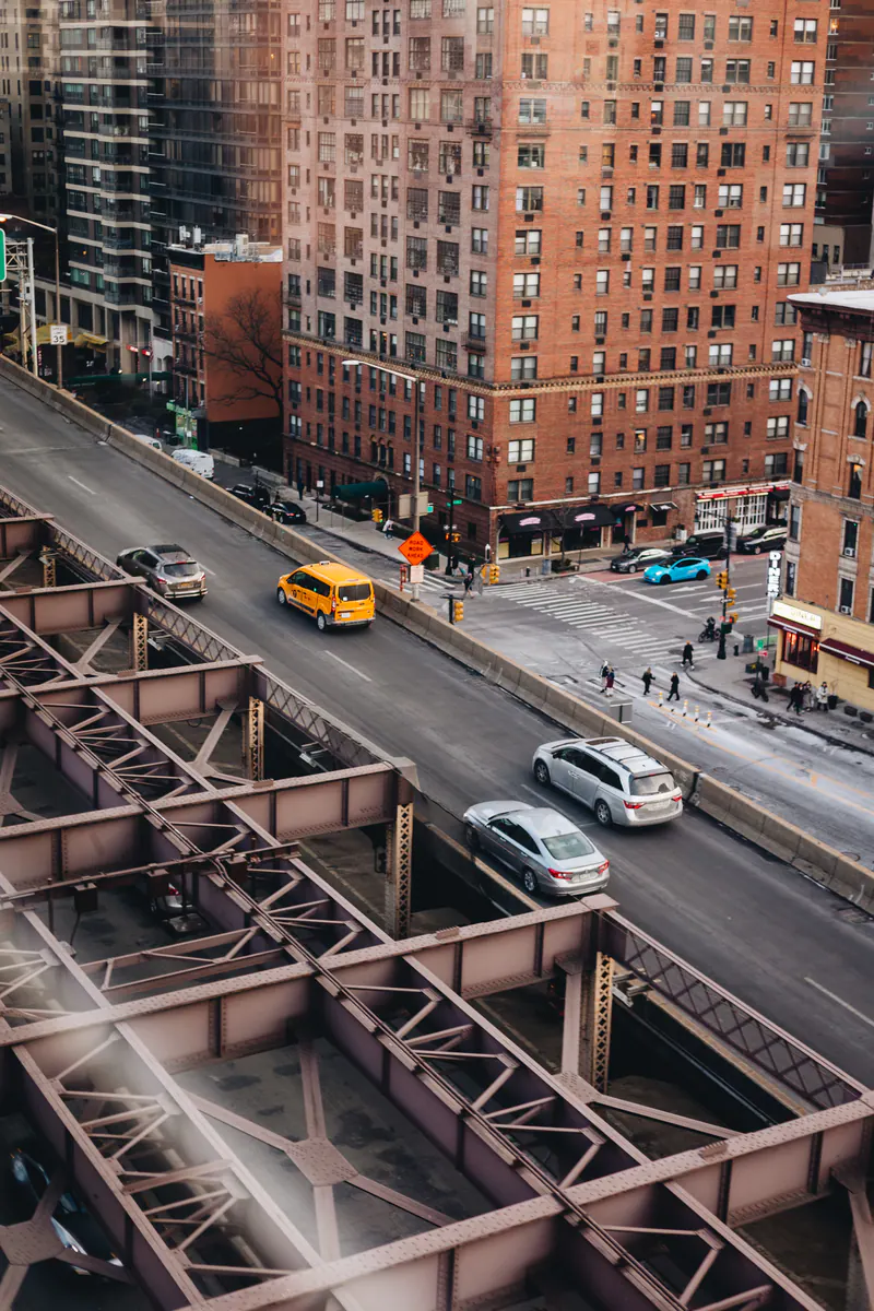 A yellow taxi on a bridge ramp beside brick apartment buildings, seen from above