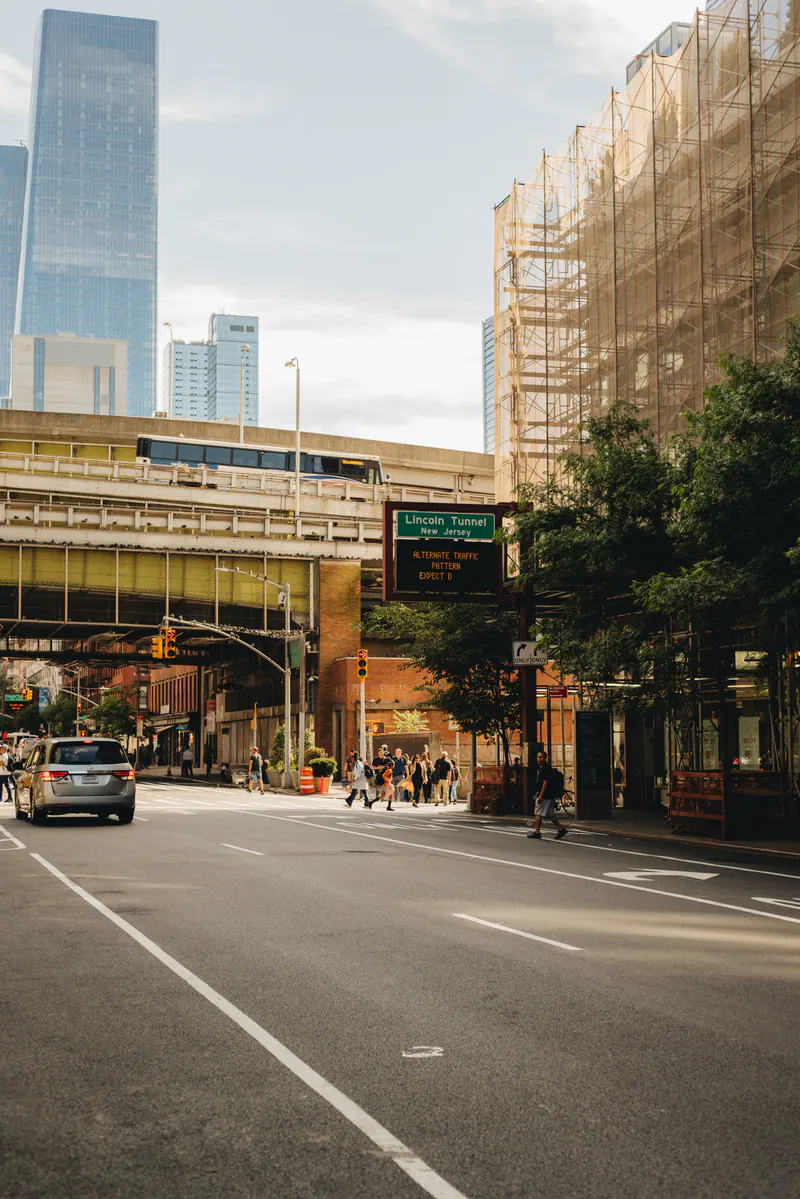 A sunlit street running towards glass towers, with an elevated road crossing it