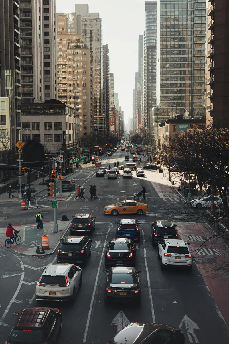 Traffic waiting on an avenue between towers, with a yellow taxi crossing